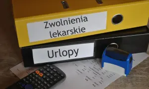 Zółty segregator "Zwolnienia lekarskie" i czarny "Urlopy" obok kalkulatora i pieczątki. Dokumenty na biurku sugerują, że kod a na zwolnieniu jest w trakcie rozliczeń.