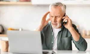 Zmartwiony mężczyzna rozmawia przez telefon, zastanawiając się, ile wynosi siła wyższa. Na stole laptop i kubek.