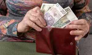 Dłoń starszej osoby wkłada polskie banknoty do brązowego portfela. To pieniądze, ich ekwiwalent.