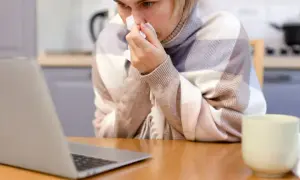 Chora kobieta z chusteczką przy nosie, owinięta kocem, sprawdza w internecie, ile wynosi zasiłek chorobowy.