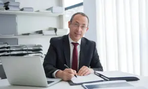 Mężczyzna w garniturze pisze coś na papierze, obok laptop i stosy dokumentów. Jego zaangażowanie w pracę jest widoczne.