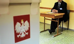 Mąż zaufania w garniturze siedzi przy biurku w lokalu wyborczym, obok urny z godłem Polski.