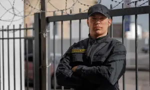 Kwalifikowany pracownik ochrony w czarnej kurtce z naszywką "SECURITY" stoi przy ogrodzeniu z drutem kolczastym.