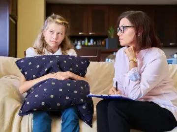 Dziewczynka z poduszką rozmawia z kobietą, która notuje. Może to być rozmowa o tym, ile zarabia kurator sądowy.