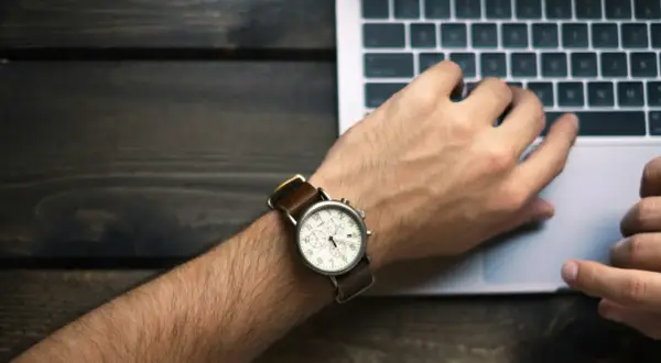 Nadgarstek z zegarkiem i dłonie na klawiaturze laptopa symbolizują nienormowany czas pracy.