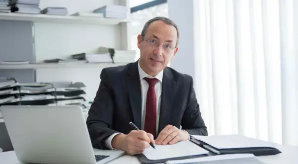 Mężczyzna w garniturze pisze coś na papierze, obok laptop i stosy dokumentów. Jego zaangażowanie w pracę jest widoczne.