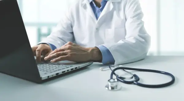 Lekarz w białym kitlu wykonuje badania wstępne do pracy, wpisując dane na laptopie. Obok leży stetoskop.