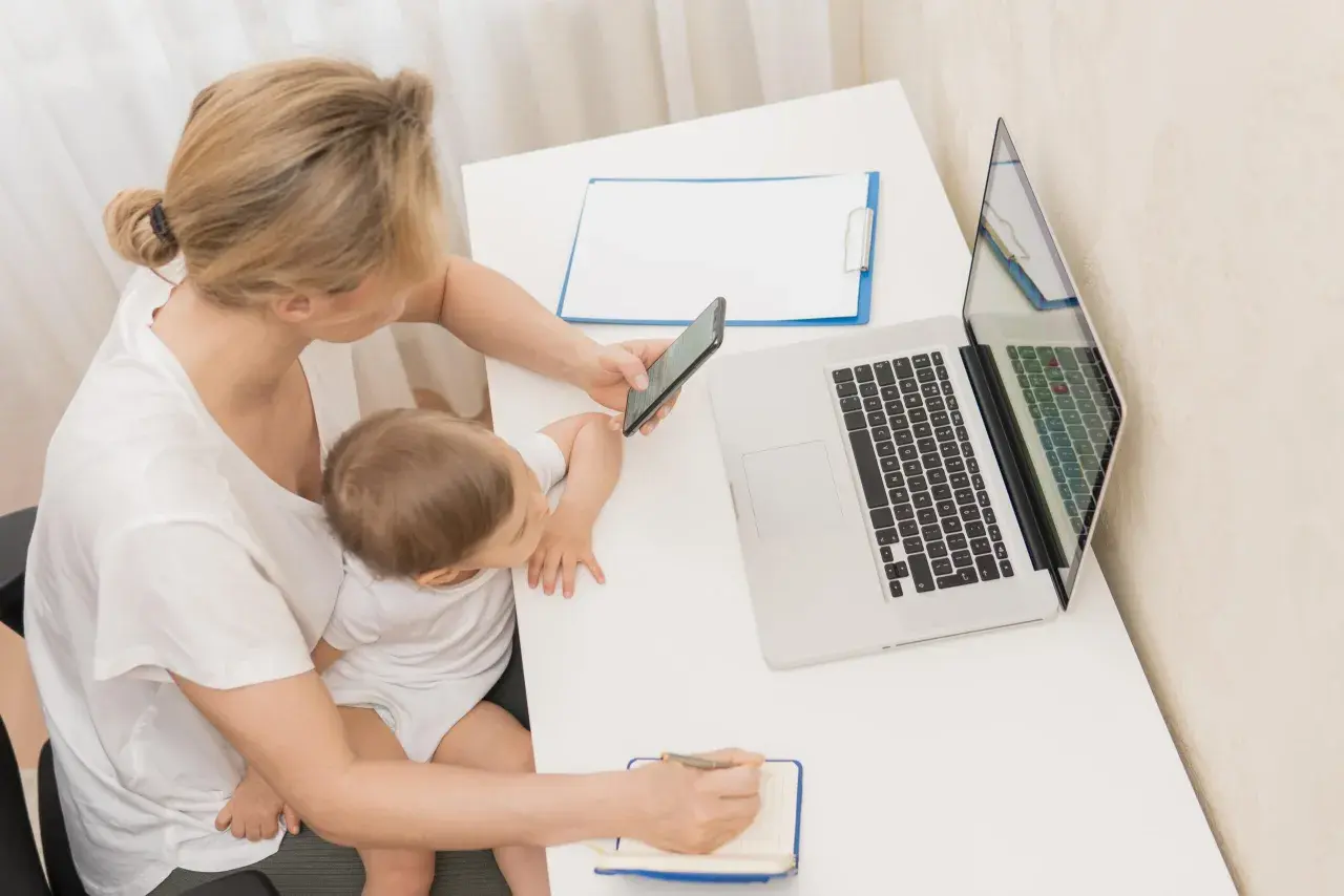 Kobieta z dzieckiem na kolanach sprawdza telefon, obok laptop i notatnik. Planuje wnioski o zasiłek macierzyński.