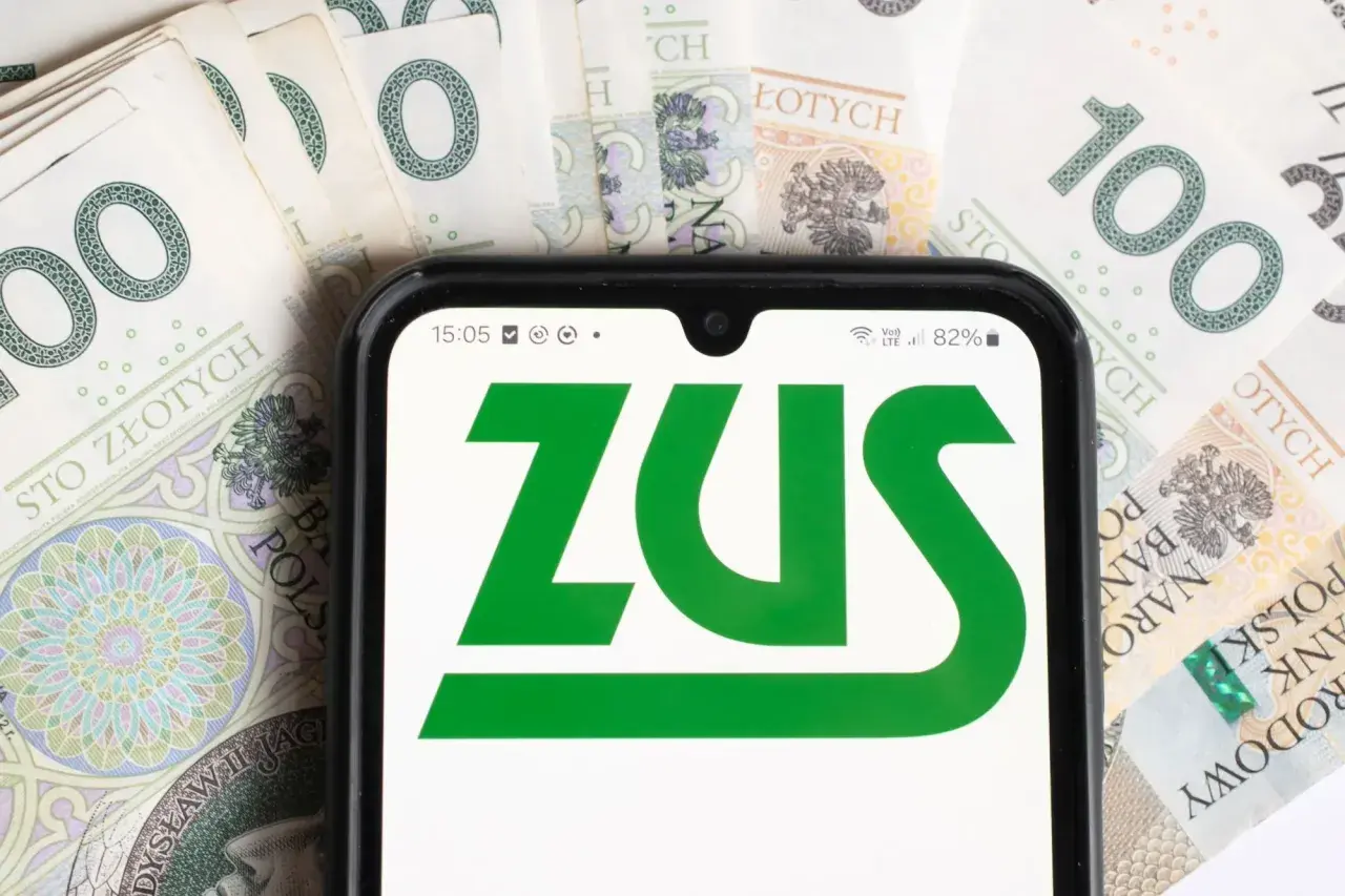 ZUS na ekranie telefonu na tle banknotów 100 zł. Sprawdź aktualną stopę procentową składek na ubezpieczenie wypadkowe.