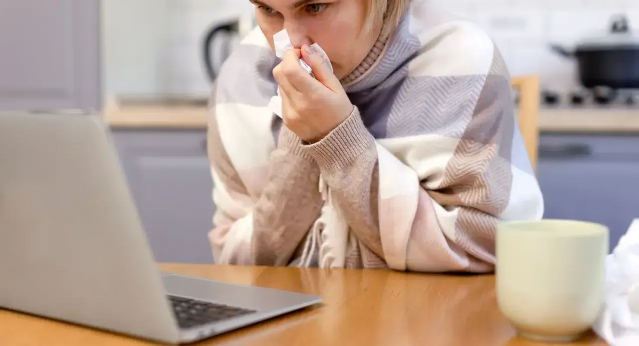 Chora kobieta z chusteczką przy nosie, owinięta kocem, sprawdza w internecie, ile wynosi zasiłek chorobowy.