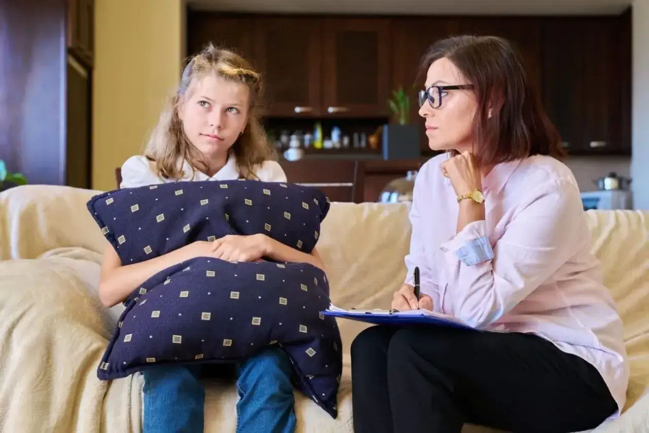 Dziewczynka z poduszką rozmawia z kobietą, która notuje. Dyskutują o tym, ile zarabia kurator sądowy.