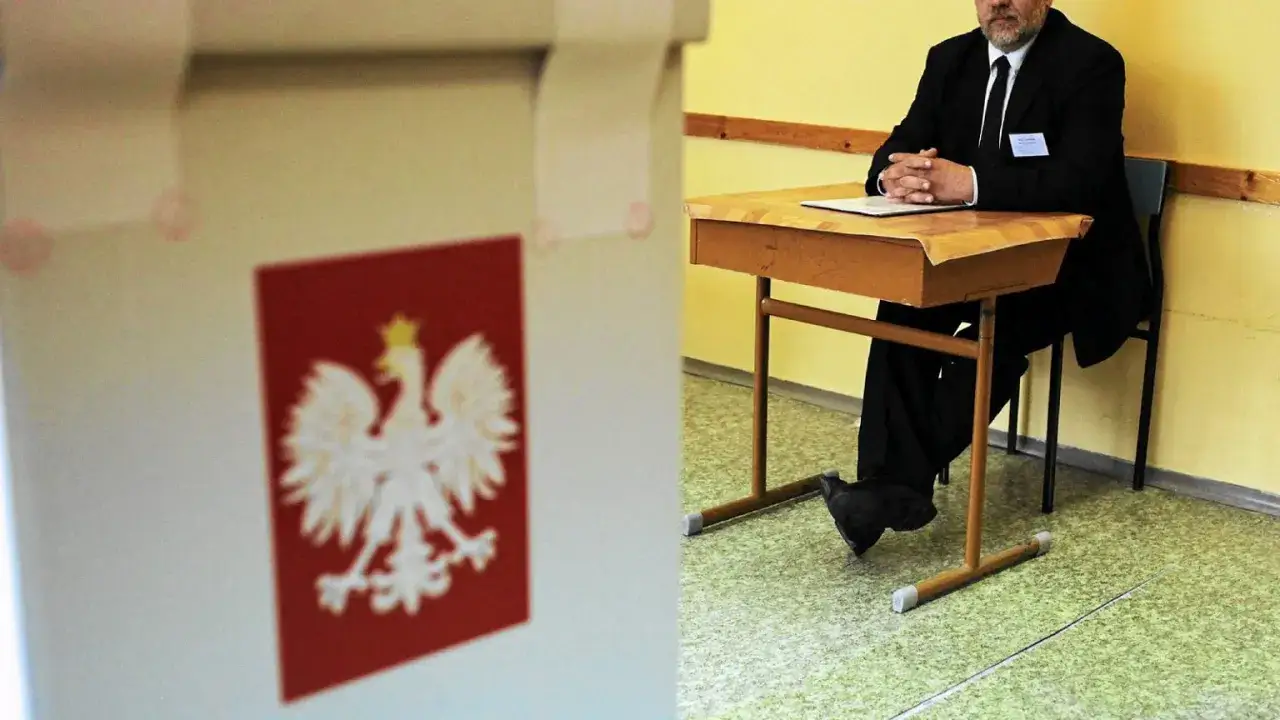 Mąż zaufania w garniturze siedzi przy biurku w lokalu wyborczym. Obok urna z godłem Polski.
