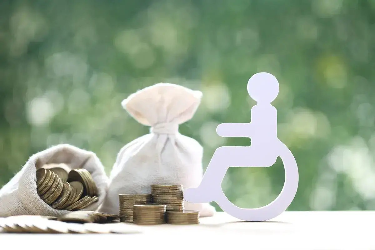Symbol osoby na wózku inwalidzkim obok worków z pieniędzmi i monetami symbolizuje wsparcie finansowe dla osób ze znacznym stopniem niepełnosprawności.
